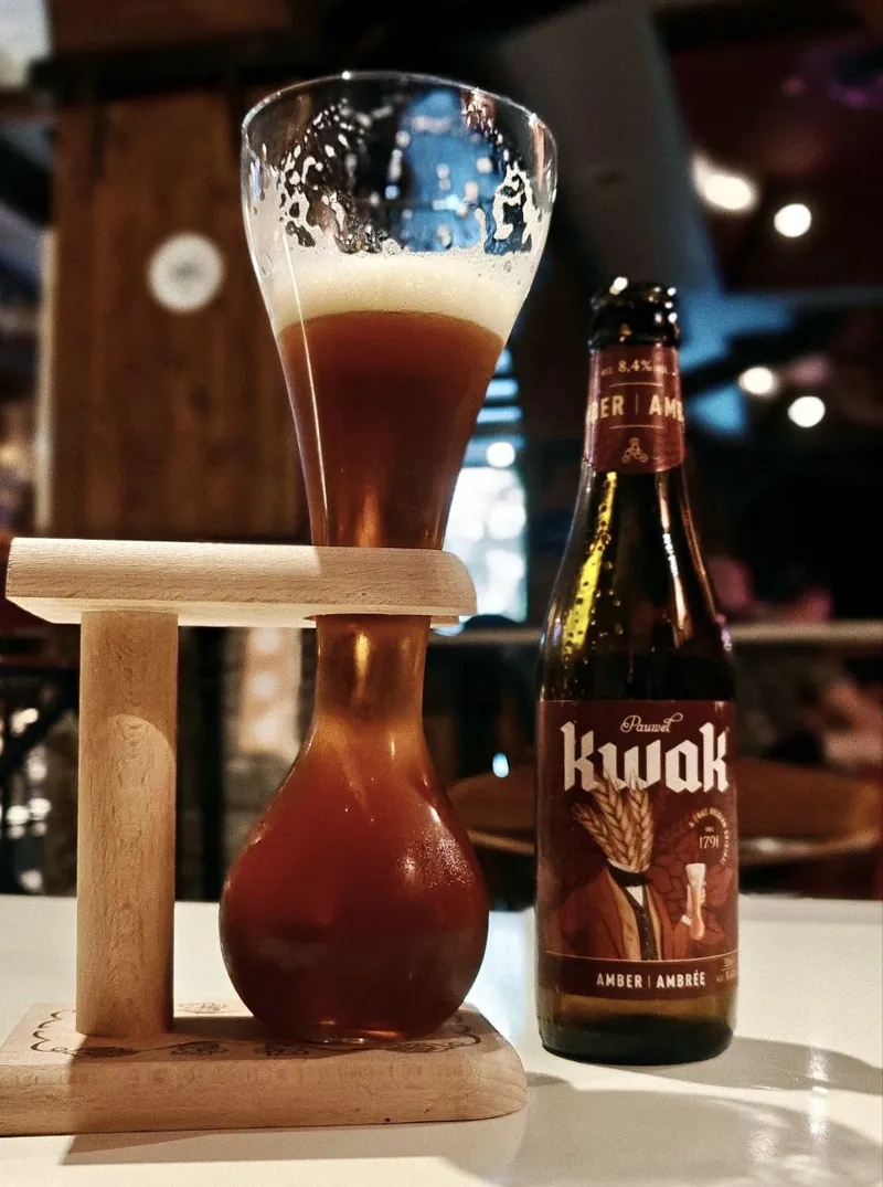 Copa de fondo redondo metida en un soporte de madera con mango, con cerveza ámbar, junto a su botella