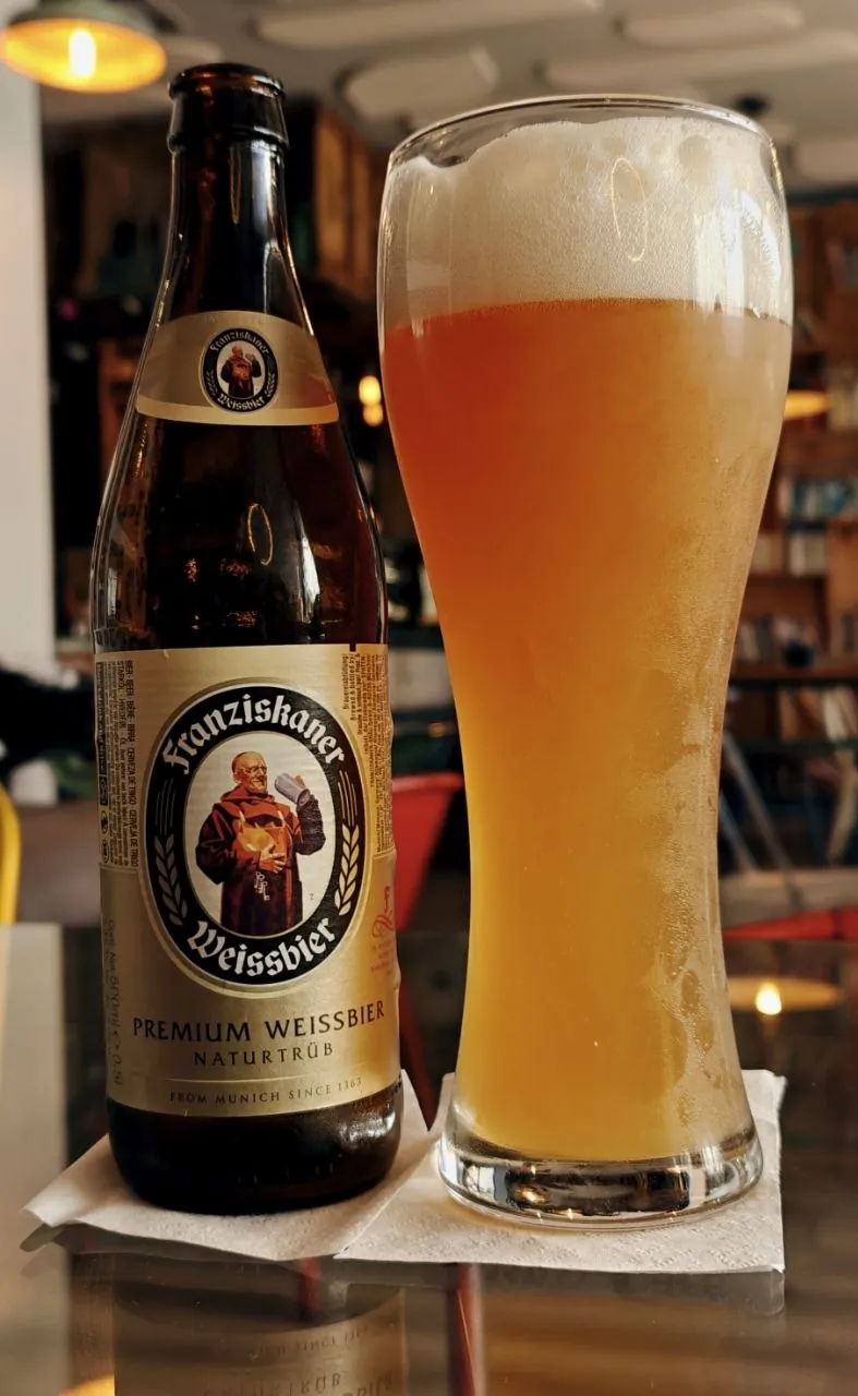 Vaso de trigo alto, estrecho abajo y más ancho arriba, con cerveza de trigo turbia y una capa alta de espuma, junto a su botella